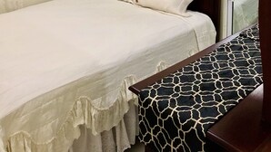 Un lit simple avec une tête de lit en bois, une table de chevet avec un coussin à motifs, un climatiseur fixé au mur et une fenêtre donnant sur un paysage agréable.