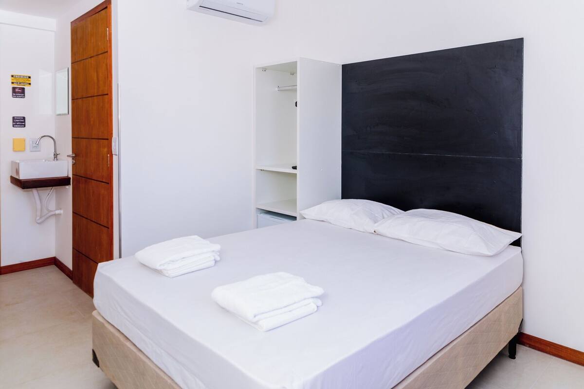 Hotellihuone, jossa on sänky, puinen vaatekaappi, valkoinen pesuallas ja musta pääty.