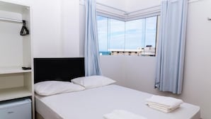 A single bed with white linens, a black headboard, and light blue curtains. There is a small white cabinet to the left and a window with curtains. - Economy HOTEL (Natal)