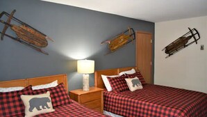 A room with two beds, each with a red and black checkered bedspread and a pillow with a bear silhouette. There is a nightstand with a lamp between the beds.