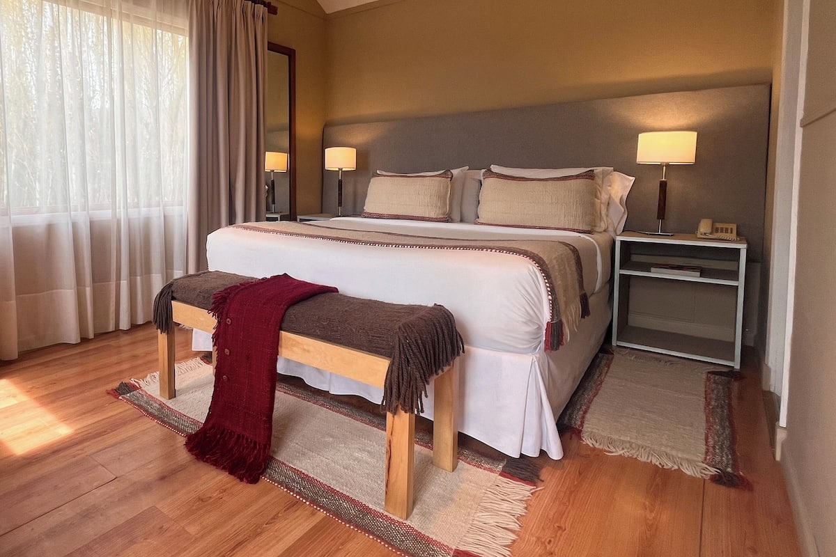 Un dormitorio con una cama grande, dos mesitas de noche con lámparas, un banco y una ventana con cortinas.