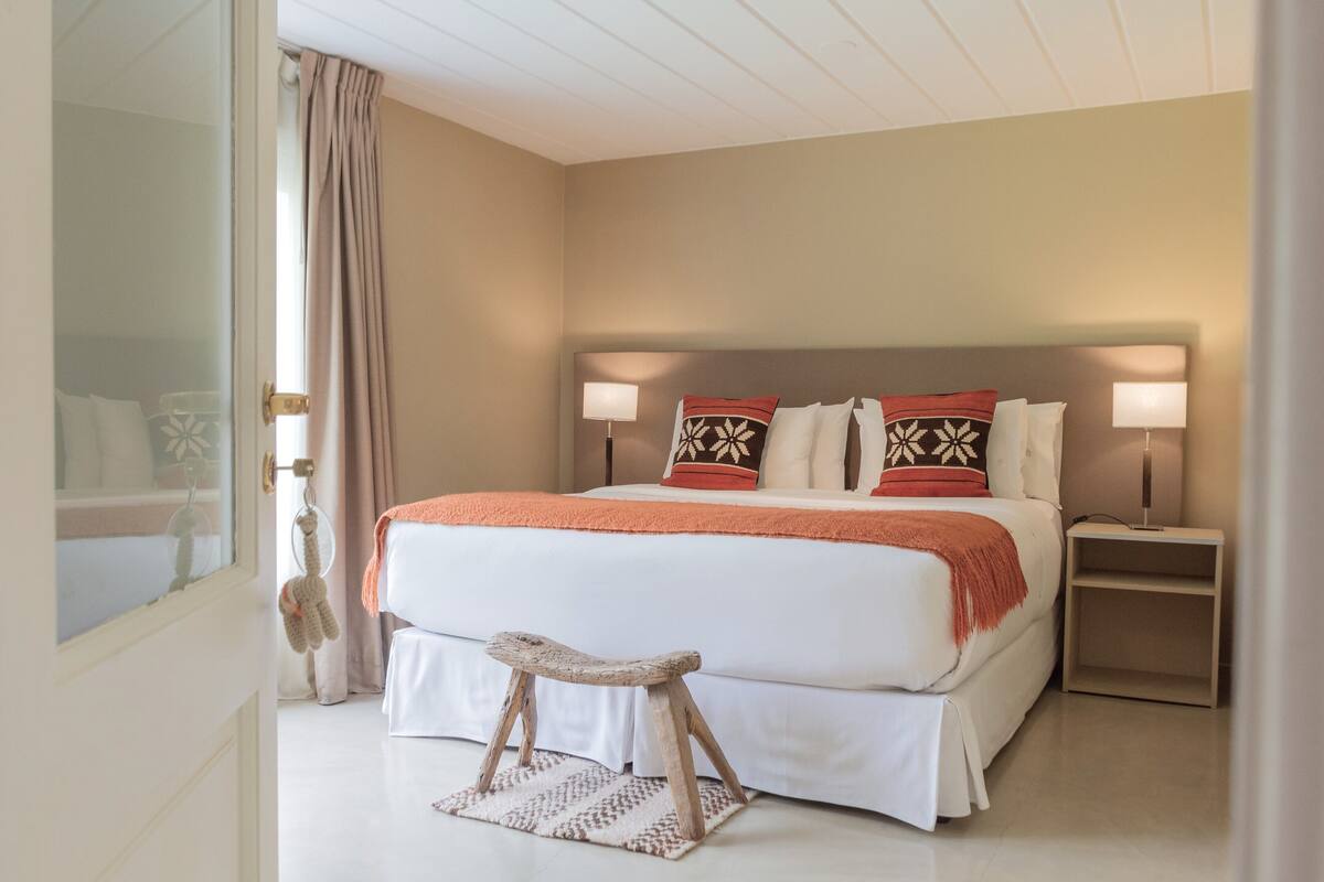 Un dormitorio con una cama grande, lámparas de noche, un taburete pequeño y una ventana con cortinas.