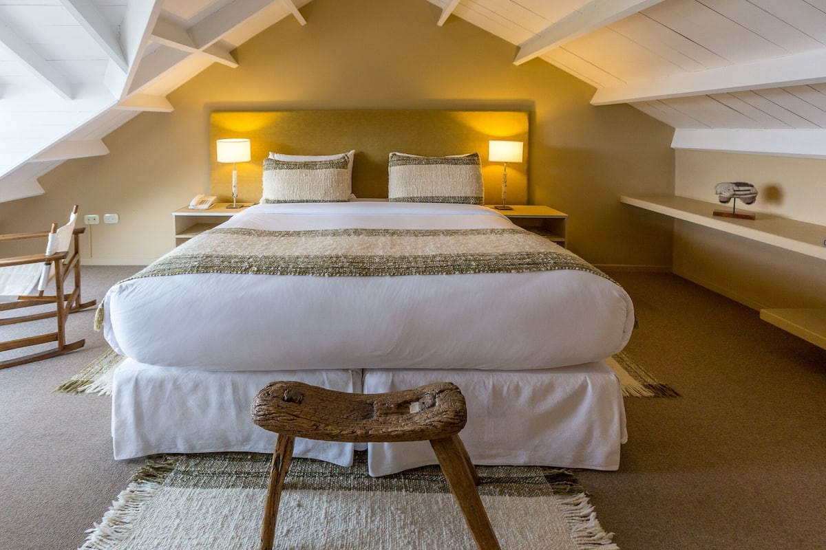 Un dormitorio con una cama grande, dos lámparas de noche, una silla de madera y un estante con un objeto decorativo.