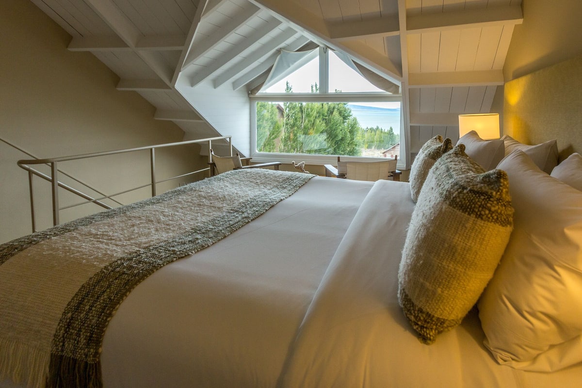 Un dormitorio con una cama grande, vistas a árboles a través de una ventana y una escalera.