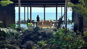 Un espacio al aire libre cubierto con vista al mar, que incluye un banco de madera, zona de estar y plantas tropicales.