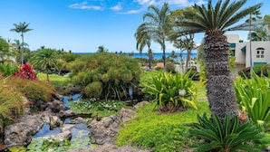 A tropical garden with a pond, palm trees, and a view of the ocean. - Palms at Wailea 703 - Modern Renovation, Prime Location, Full AC, Close to Pool! (Kihei)