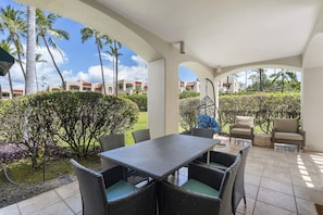 A covered outdoor patio with a table and chairs, surrounded by tropical plants and palm trees. - Palms at Wailea 703 - Modern Renovation, Prime Location, Full AC, Close to Pool! (Kihei)