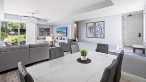A modern living room with a large sofa, dining table, and chairs. There is a TV mounted on the wall and a ceiling fan. - Palms at Wailea 703 - Modern Renovation, Prime Location, Full AC, Close to Pool! (Kihei)