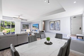 Ein modernes Wohnzimmer mit einer großen Couch, einem Esstisch und Stühlen. Ein Fernseher ist an der Wand befestigt und ein Deckenventilator vorhanden.