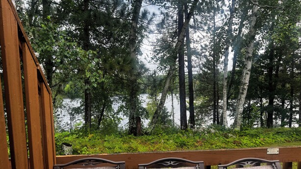 A wooden deck with three chairs facing a forested area and a body of water.