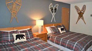 A room with two beds, each with plaid bedding and a bear pillow. There are wall-mounted decorative items and a nightstand with a lamp.
