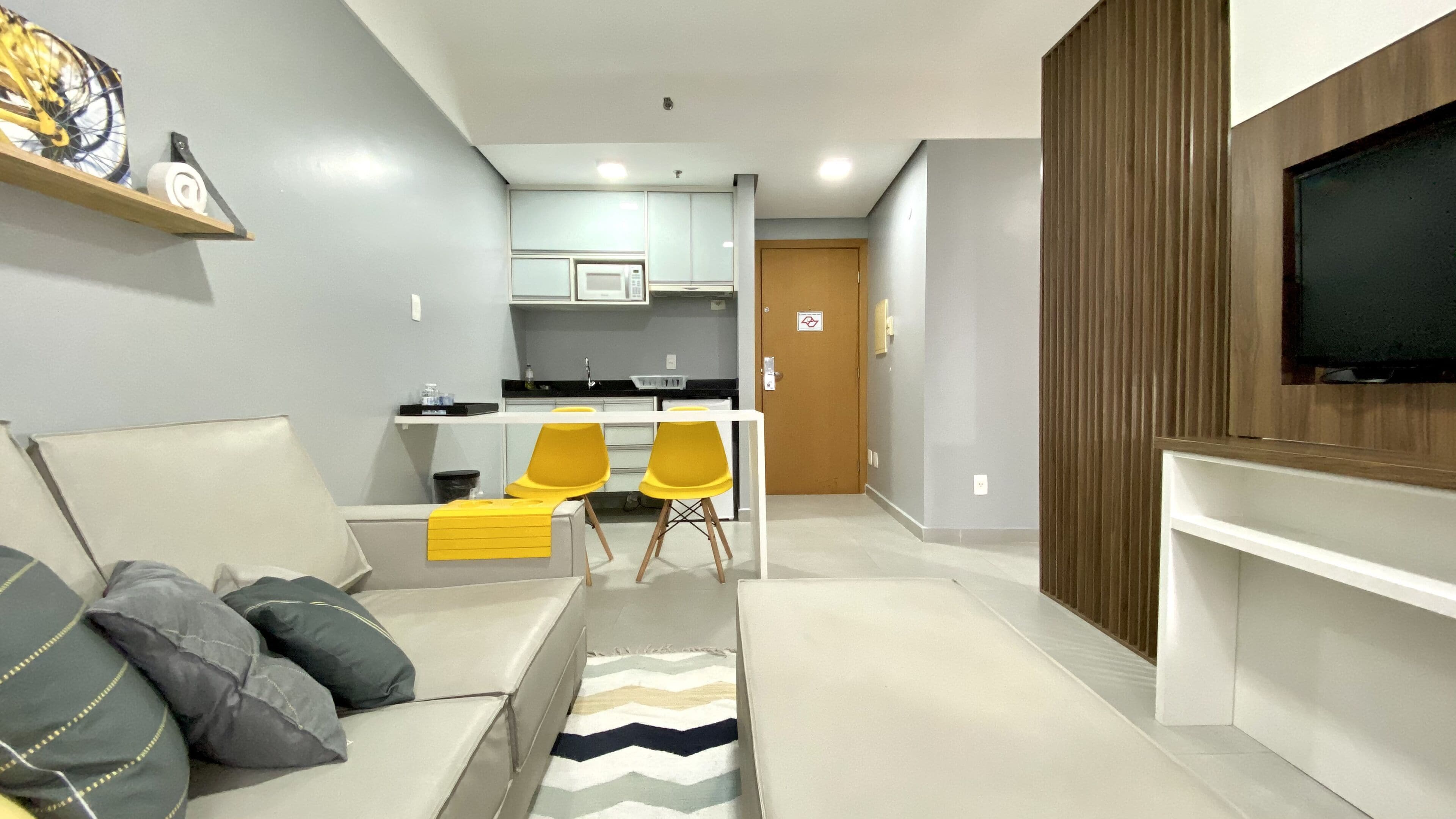 Ein modernes Wohnzimmer mit einer Couch, einem Couchtisch und einem an der Wand montierten Fernseher. Ein Küchenbereich mit einer Mikrowelle und einem Spülbecken ist ebenfalls zu sehen.