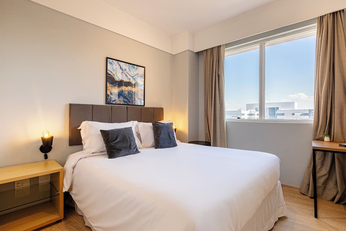 Hotellihuone, jossa on suuri sänky, yöpöytä, yövalaisin, ikkuna verhoilla ja näkymä kaupunkiin.