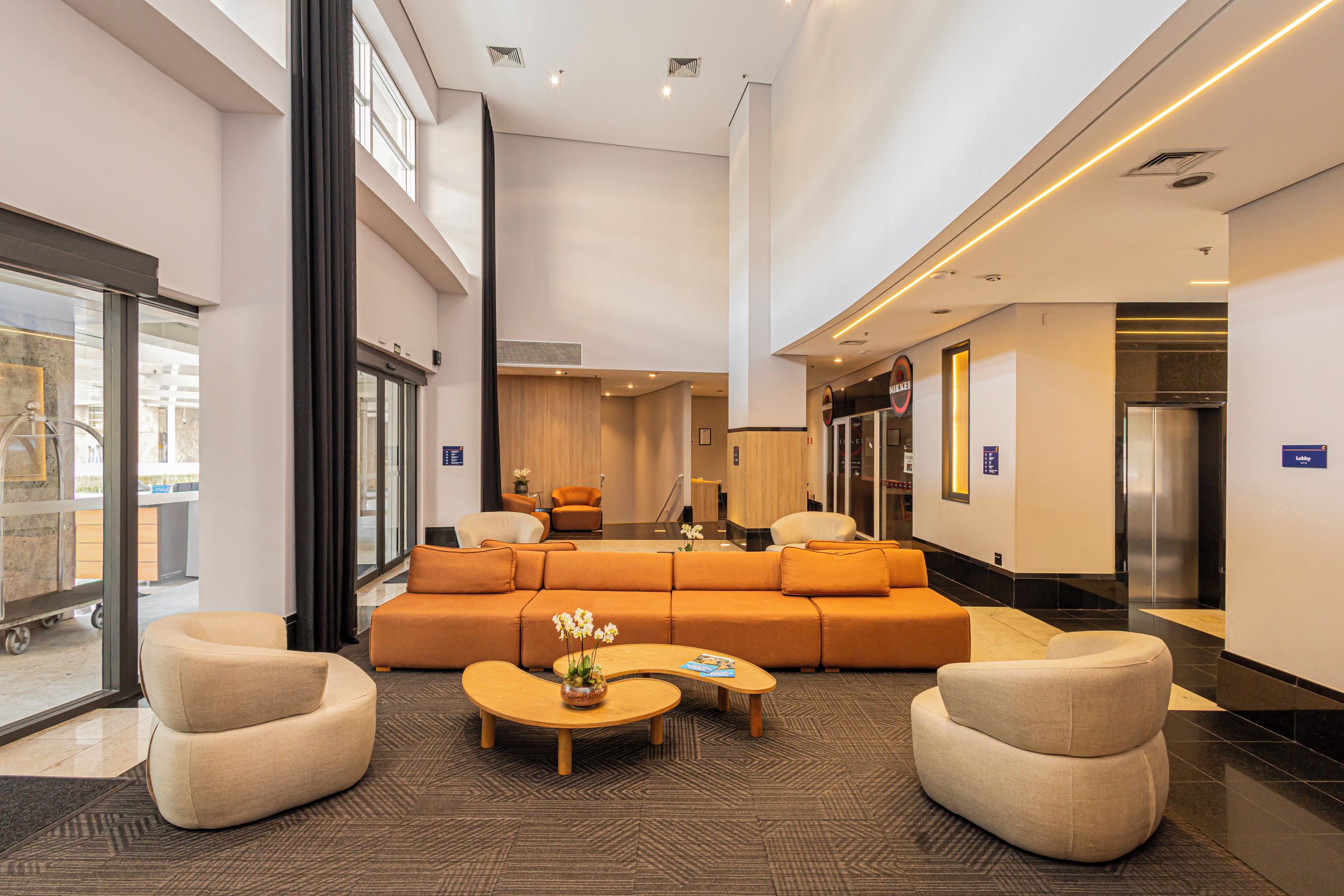 Eine großzügige Lobby mit einer großen orangefarbenen Couch, zwei Sesseln und einem Couchtisch. Dort befindet sich eine Rezeption, eine Verkaufsmaschine und ein Aufzug.