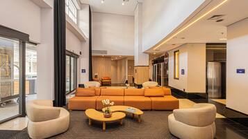 Eine großzügige Lobby mit einer großen orangefarbenen Couch, zwei Sesseln und einem Couchtisch. Dort befindet sich eine Rezeption, eine Verkaufsmaschine und ein Aufzug.