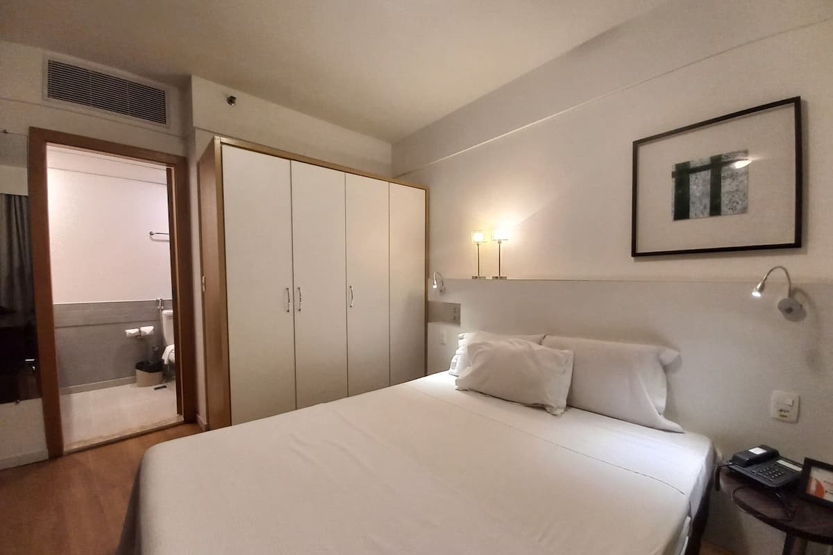 Hotellihuone, jossa on sänky, vaatekaappi, yöpöytä, jossa on puhelin, ja seinällä kehystetty kuva.