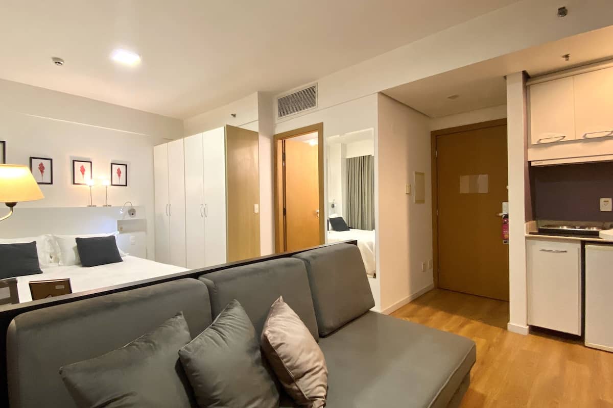 Moderni hotellihuone, jossa on sänky, harmaa sohva, lamppuvarusteinen työpöytä, pieni keittiö ja kylpyhuone, joka näkyy avoimesta ovesta.