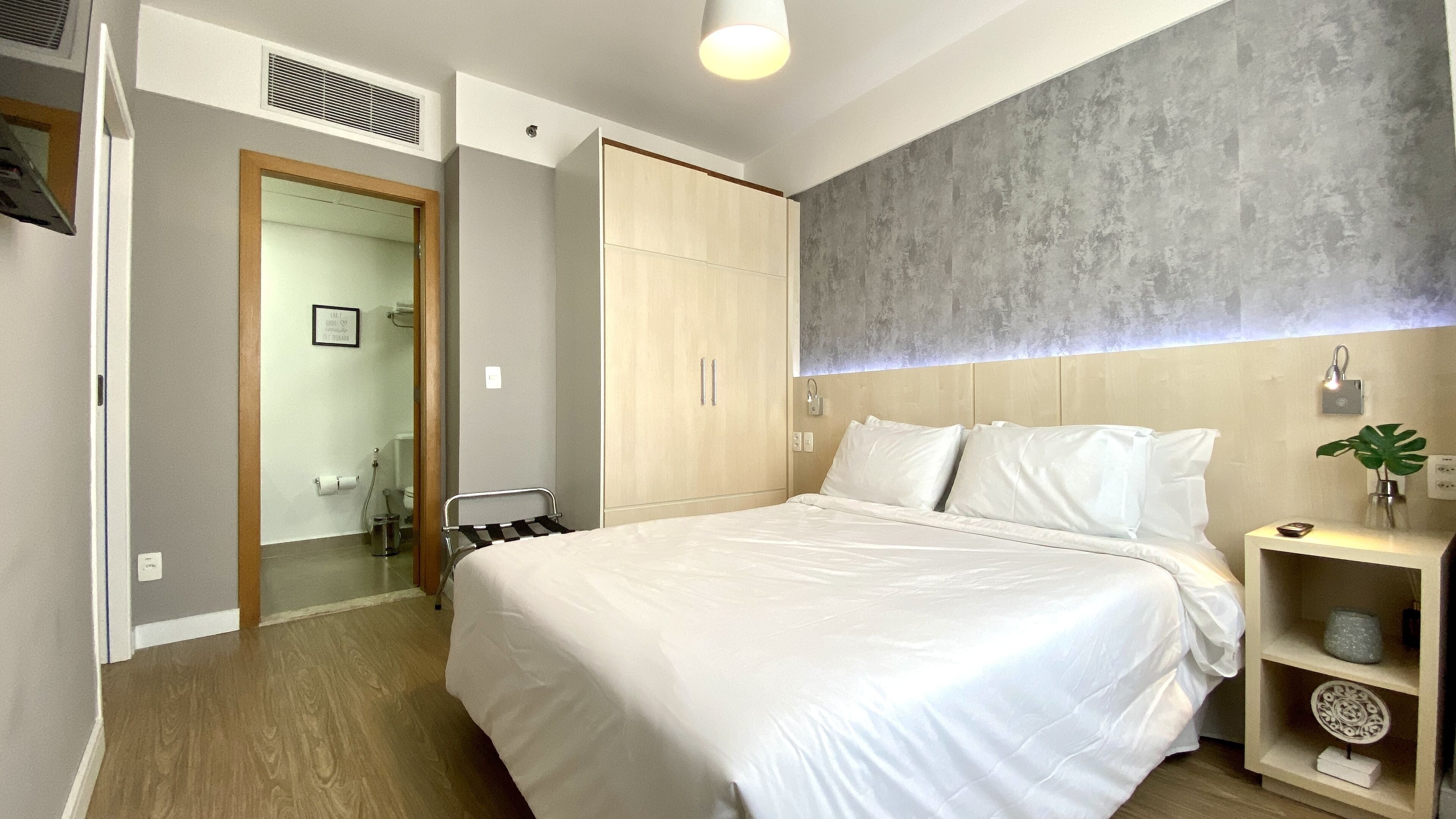 A modern bedroom with a large bed, a wooden wardrobe, a bedside table with a plant, and a grey textured wall.