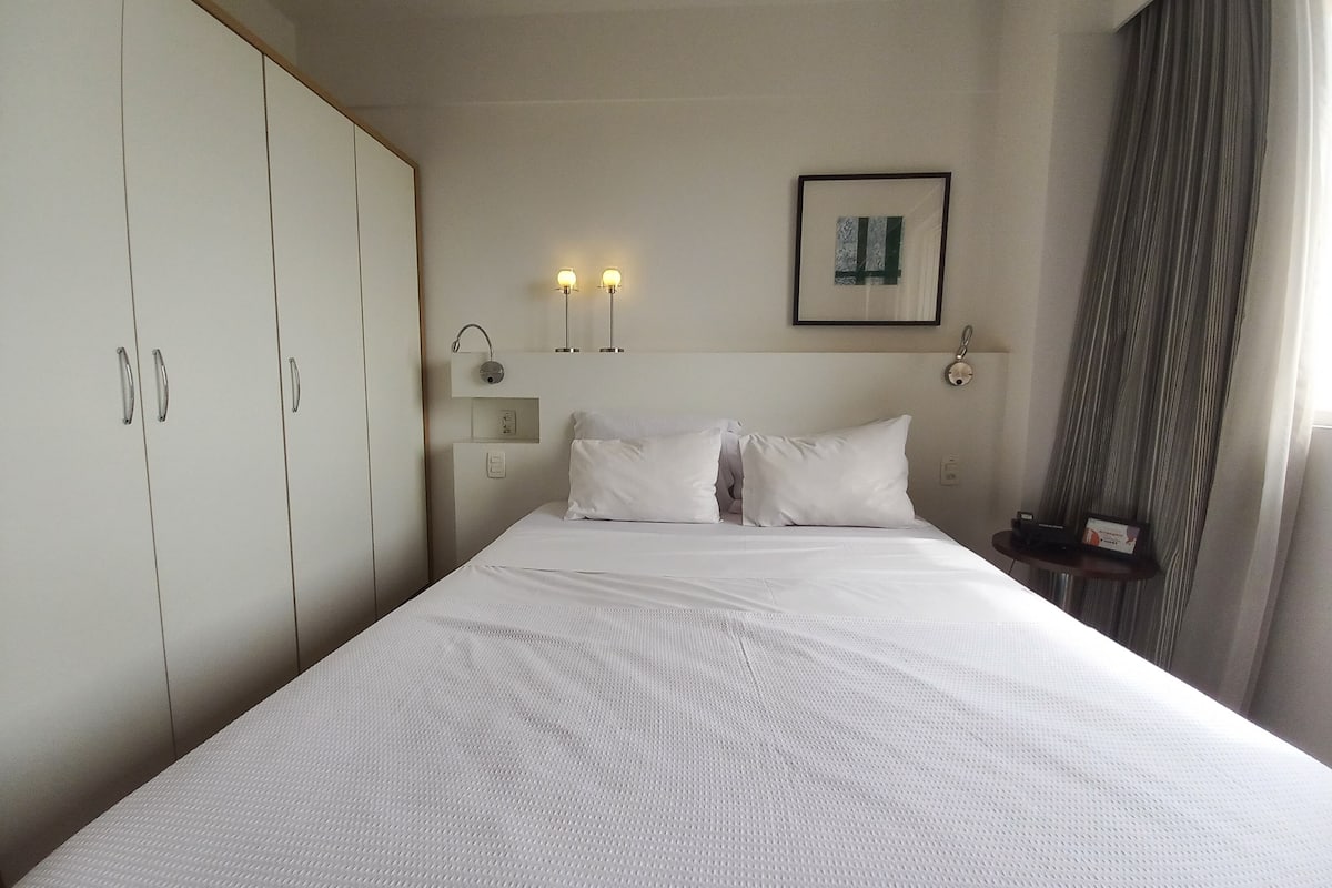 Hotellihuone, jossa on suuri sänky, vaatekaappi, yöpöytä lampun kanssa ja seinällä oleva kehystetty kuva.