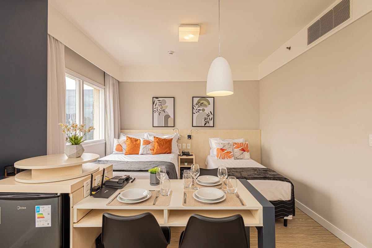 Moderni hotellihuone, jossa on ruokailutila, kaksi sänkyä, työpöytä ja jääkaappi.