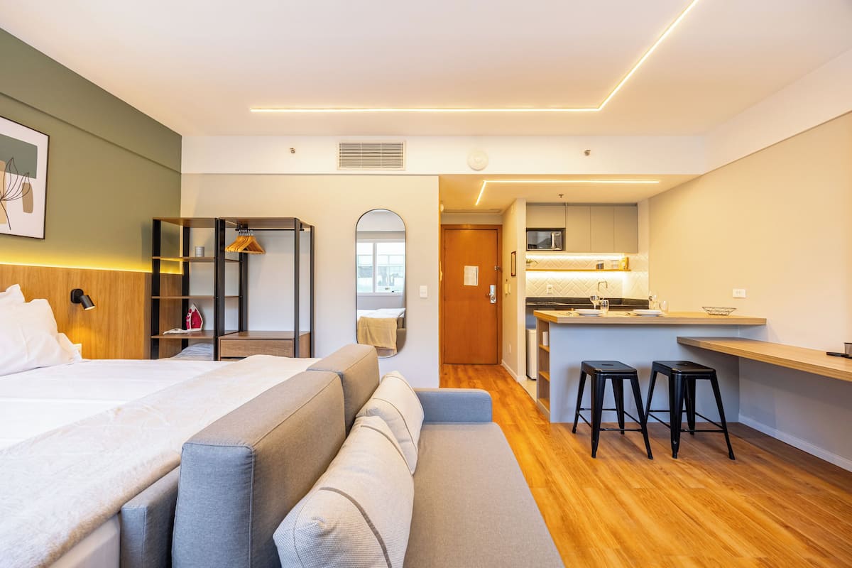 Moderni hotellihuone, jossa on suuri sänky, sohva, pieni keittiö ja kylpyhuone, joka näkyy holvikaaren läpi.