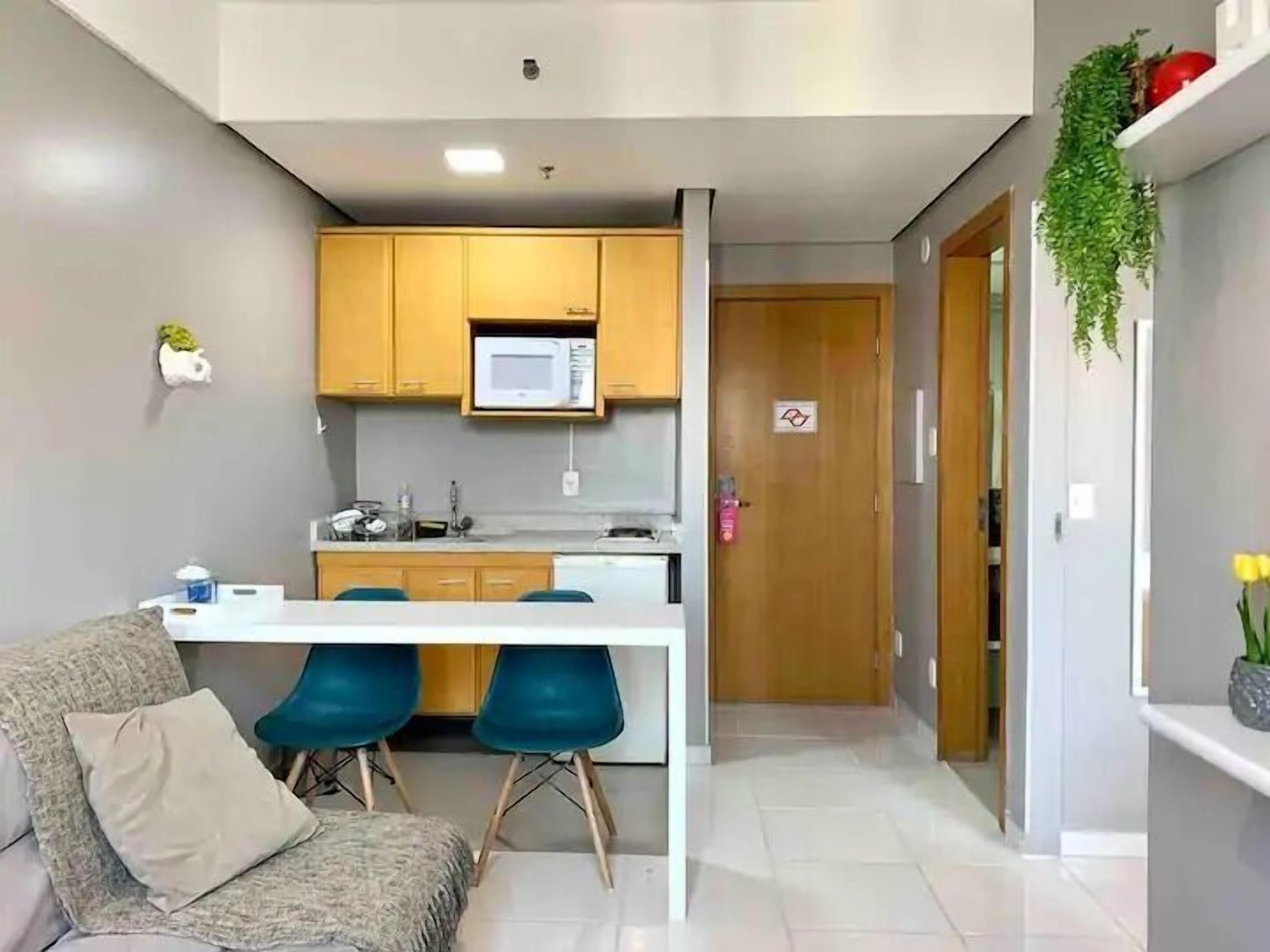 A compact kitchen with a microwave, a sink, and a countertop. There is a sofa with a cushion and two blue chairs. A door leads to another room.