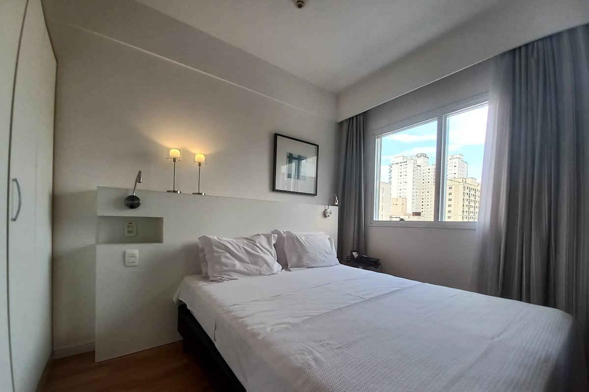 Hotellihuone, jossa on sänky, yöpöytä, seinään kiinnitetty lamppu, ikkuna verhoilla ja näkymä rakennuksiin.
