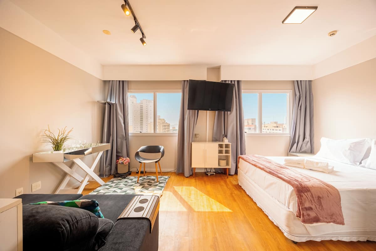 Moderni hotellihuone, jossa on sänky, työpöytä tietokoneella, tuoli ja näkymä kaupunkiin.