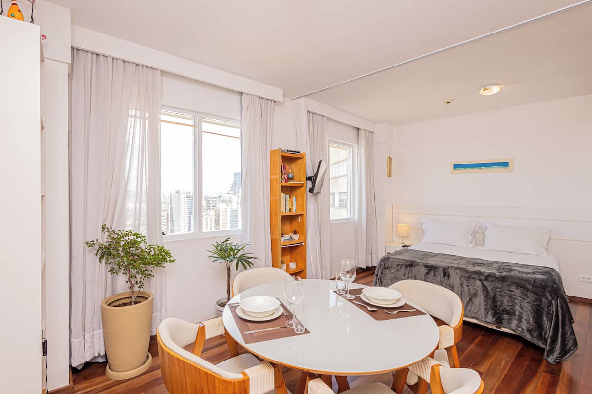 Moderni hotellihuone, jossa on sänky, kahdelle katettu ruokapöytä, kirjahylly ja suuri ikkuna, jossa on verhot.