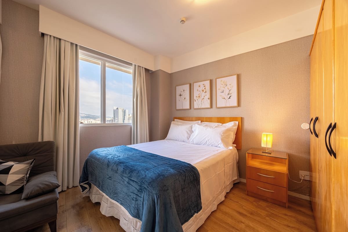 Hotellihuone, jossa on sänky, yöpöytä, vaatekaappi, ikkuna verhoilla ja näkymä kaupunkiin.