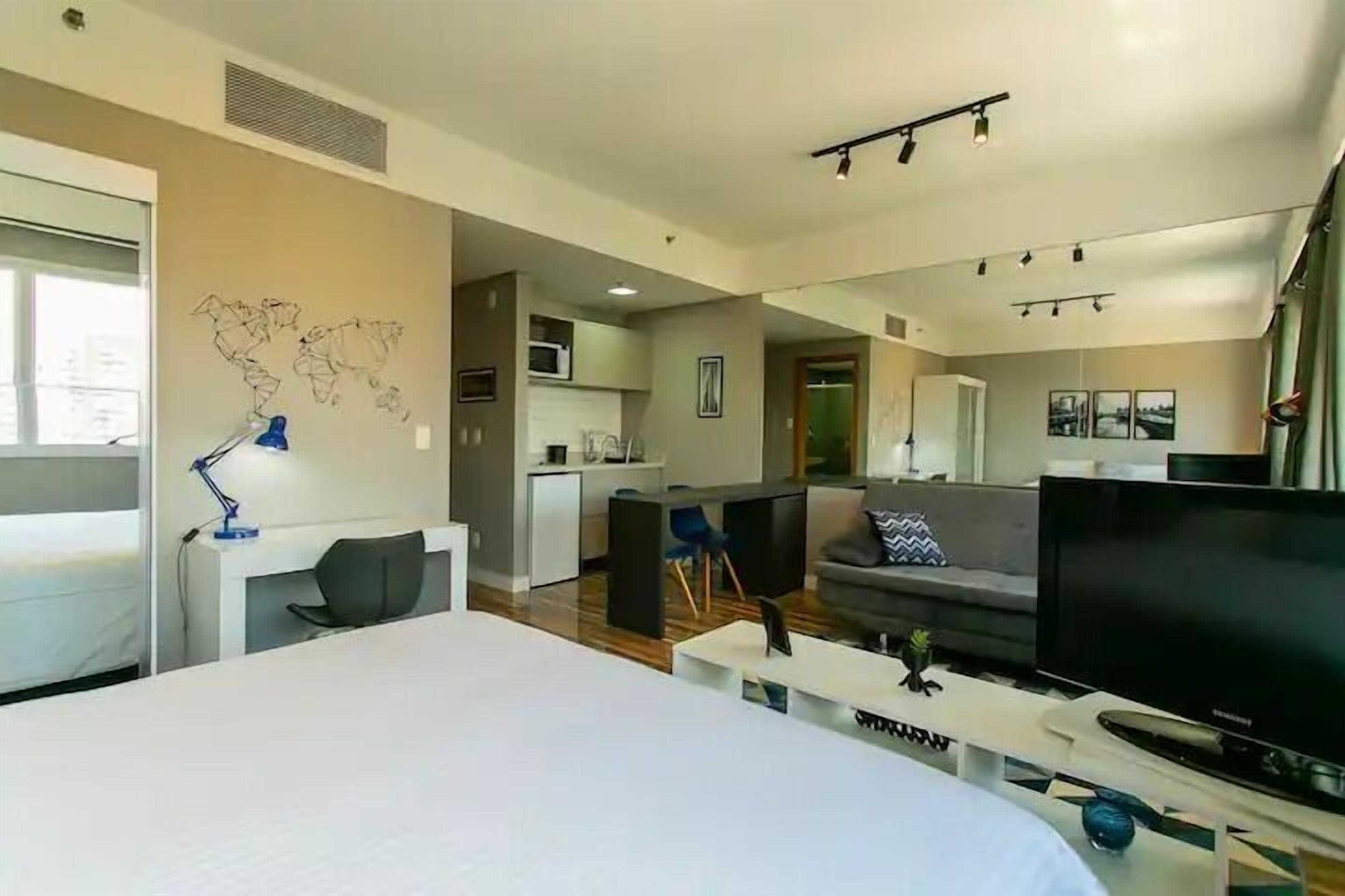 A modern hotel room with a large bed, a desk with a lamp, a TV, and a kitchenette in the background.