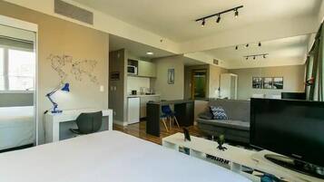 A modern hotel room with a large bed, a desk with a lamp, a TV, and a kitchenette in the background.