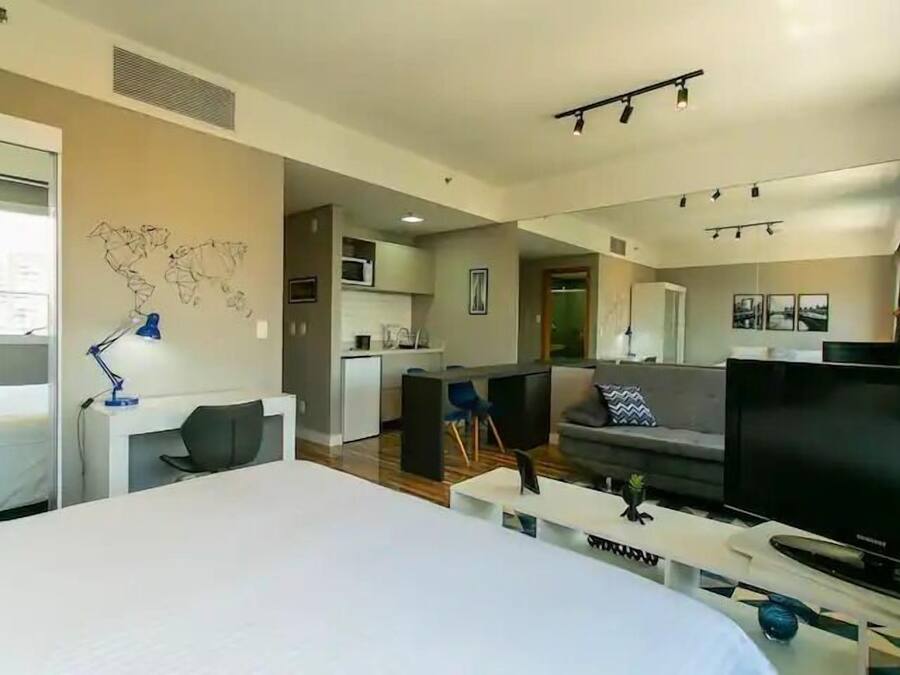 A modern hotel room with a large bed, a desk with a lamp, a TV, and a kitchenette in the background.