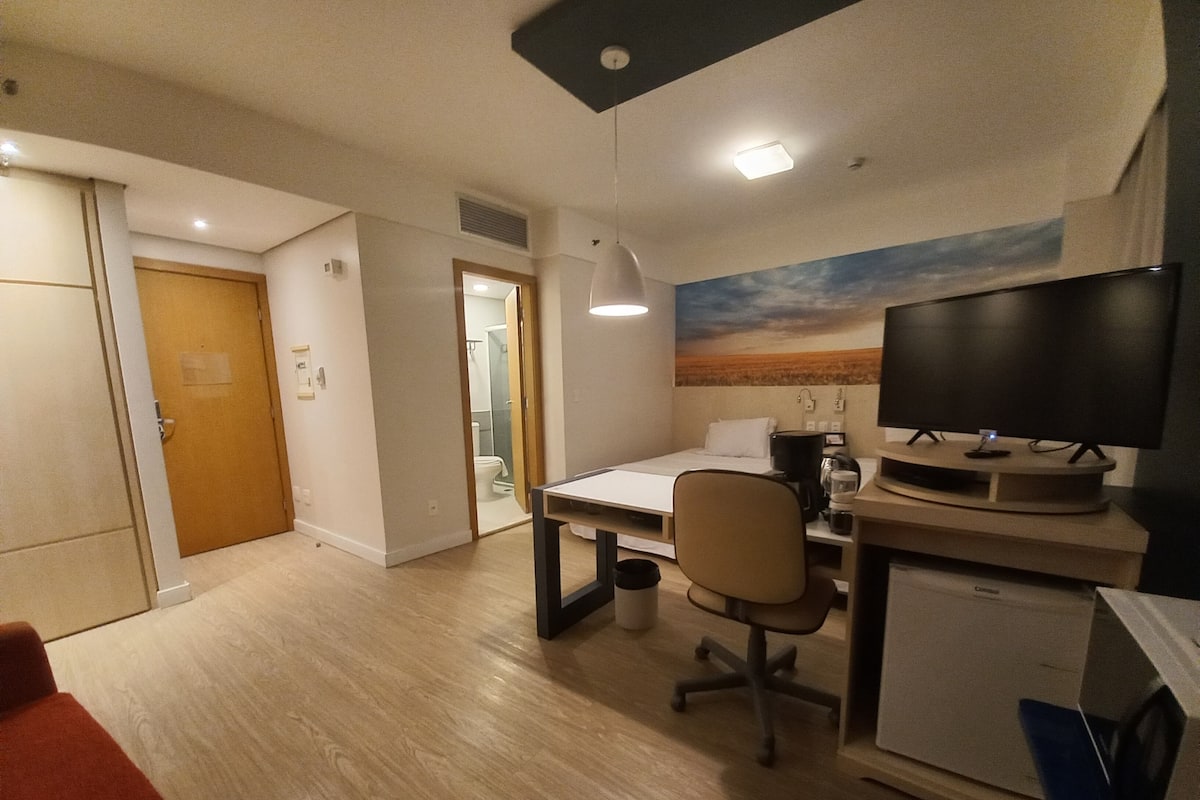 Hotellihuone, jossa on sänky, työpöytä, tuoli, televisio ja taulu seinällä.