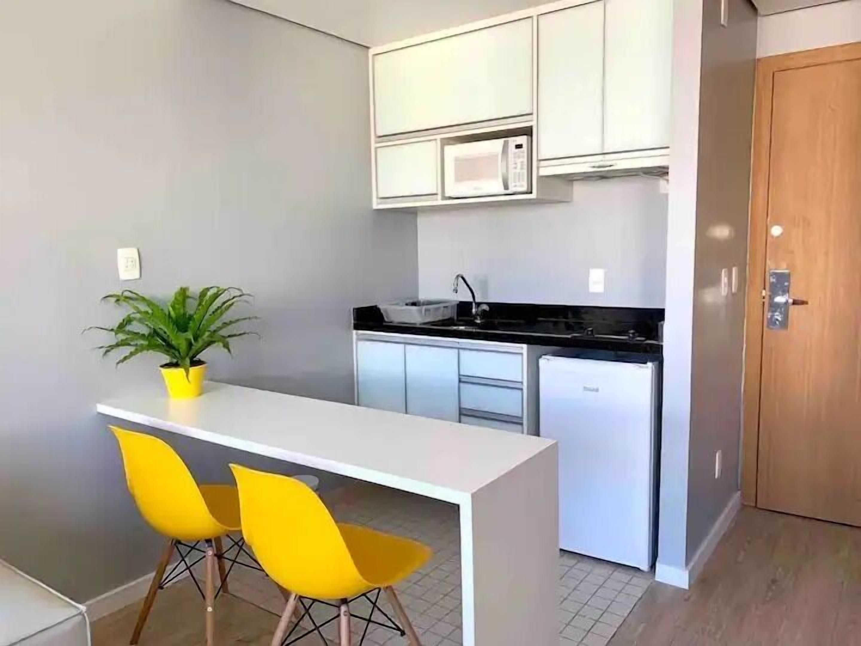 A compact kitchen with white cabinets, a microwave, and a sink. There is a small dining area with a white table and two yellow chairs. A potted plant is on the table.