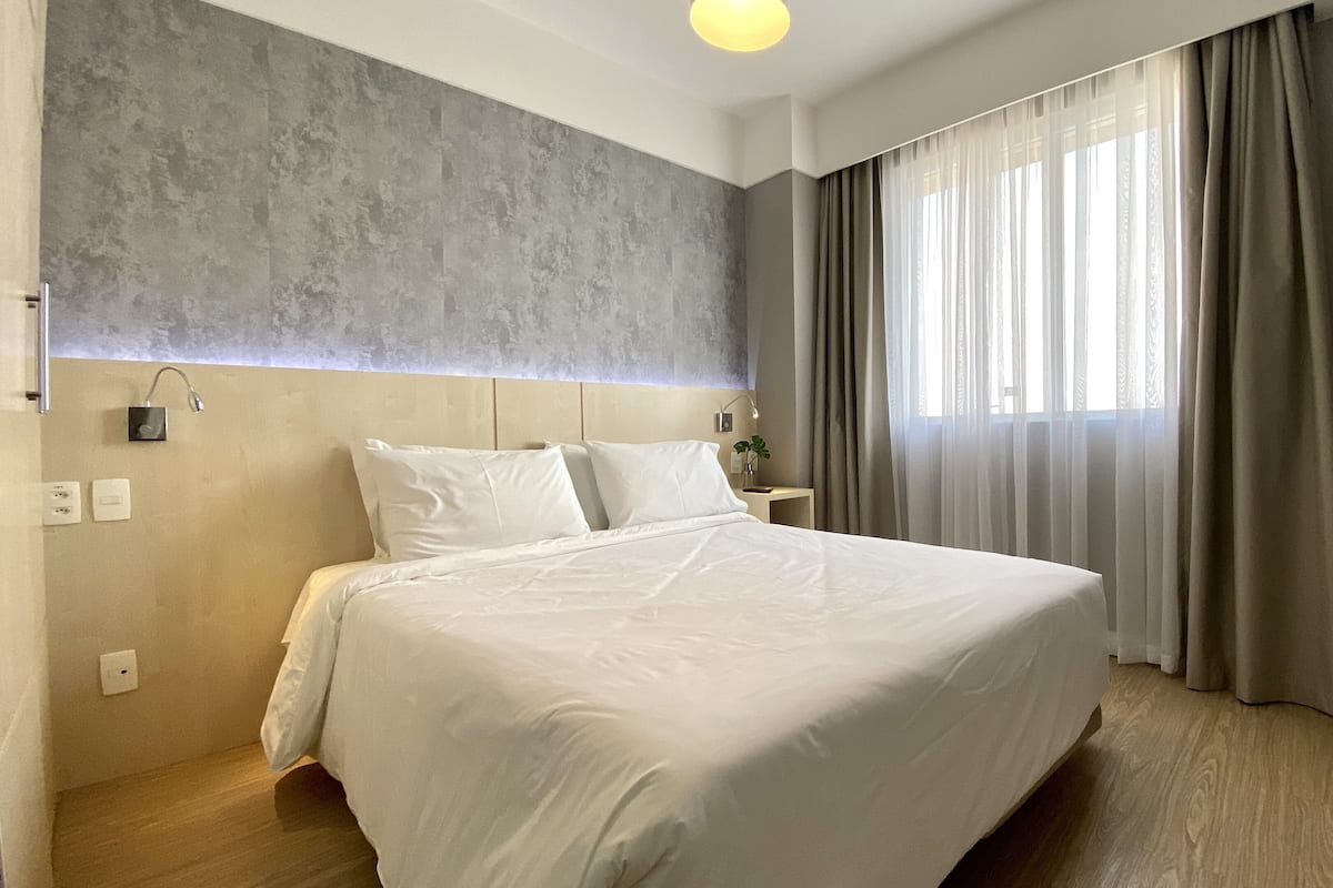 Moderni hotellihuone, jossa on suuri sänky, kuvioitu pääty, yöpöytä lampulla ja ikkuna, jossa on verhot.
