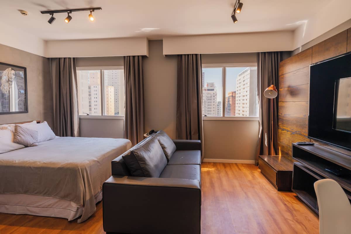 Moderni hotellihuone, jossa on sänky, sohva, televisio ja suuret ikkunat, joista avautuu näkymä kaupunkiin.