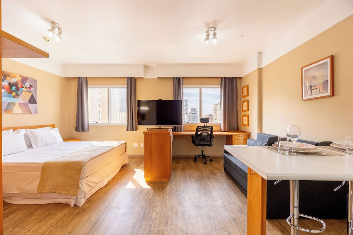 Hotellihuone, jossa on sänky, työpöytä, tuoli, televisio sekä ruokailutila, jossa on pöytä ja tuoleja.