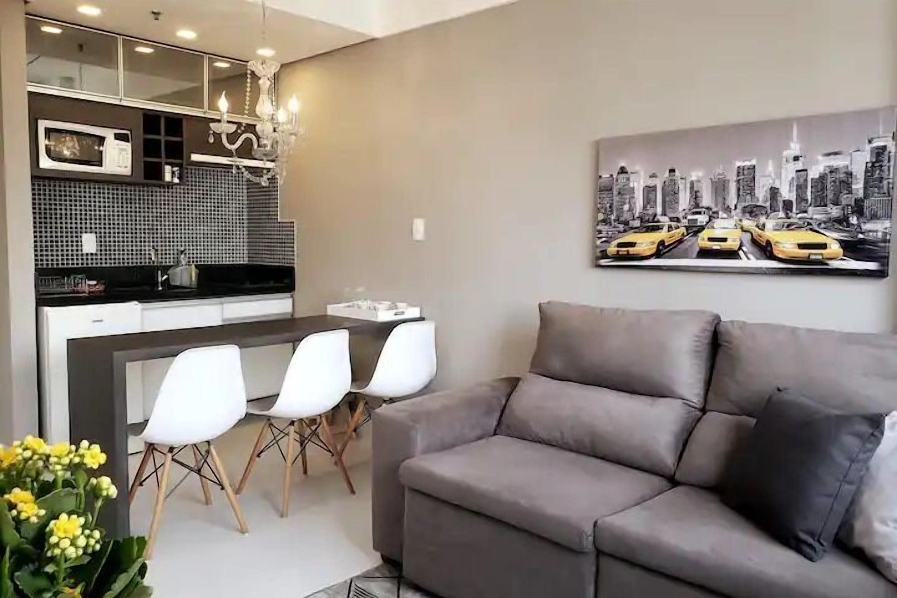 Un salon moderne avec un canapé gris, un coin repas avec des chaises blanches, une cuisine avec un plan de travail noir et un tableau représentant un paysage urbain accroché au mur.