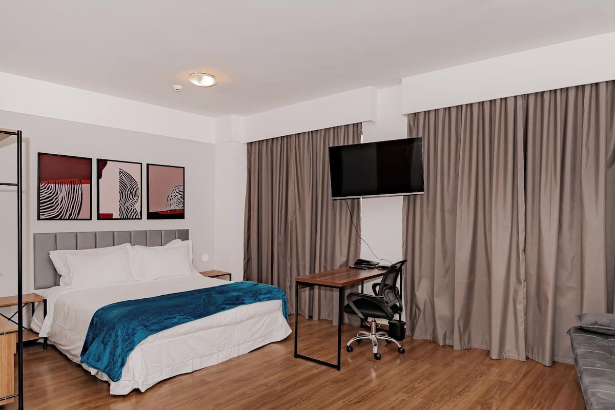 Moderni hotellihuone, jossa on suuri sänky, työpöytä, tuoli, taulutelevisio ja suuri ikkuna, jossa on verhot.