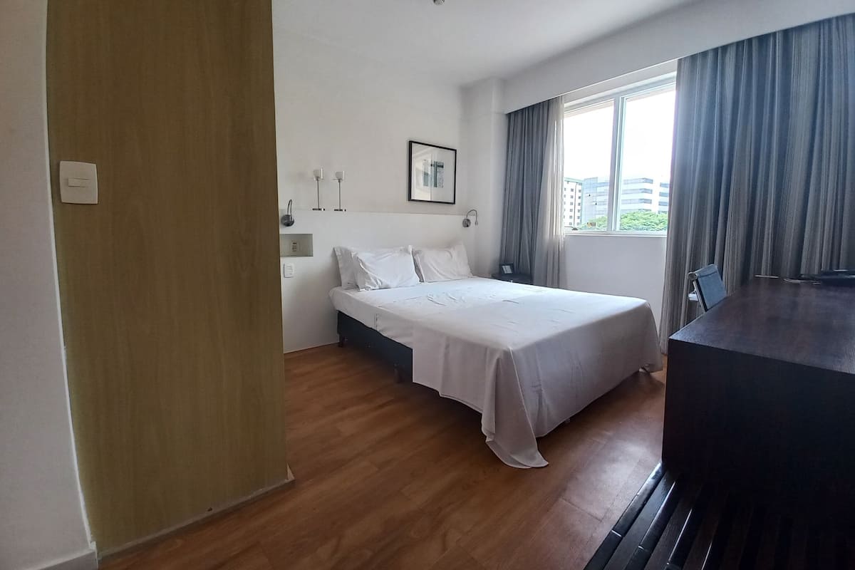 Hotellihuone, jossa on sänky, yöpöytä ja ikkuna, jossa on verhot.