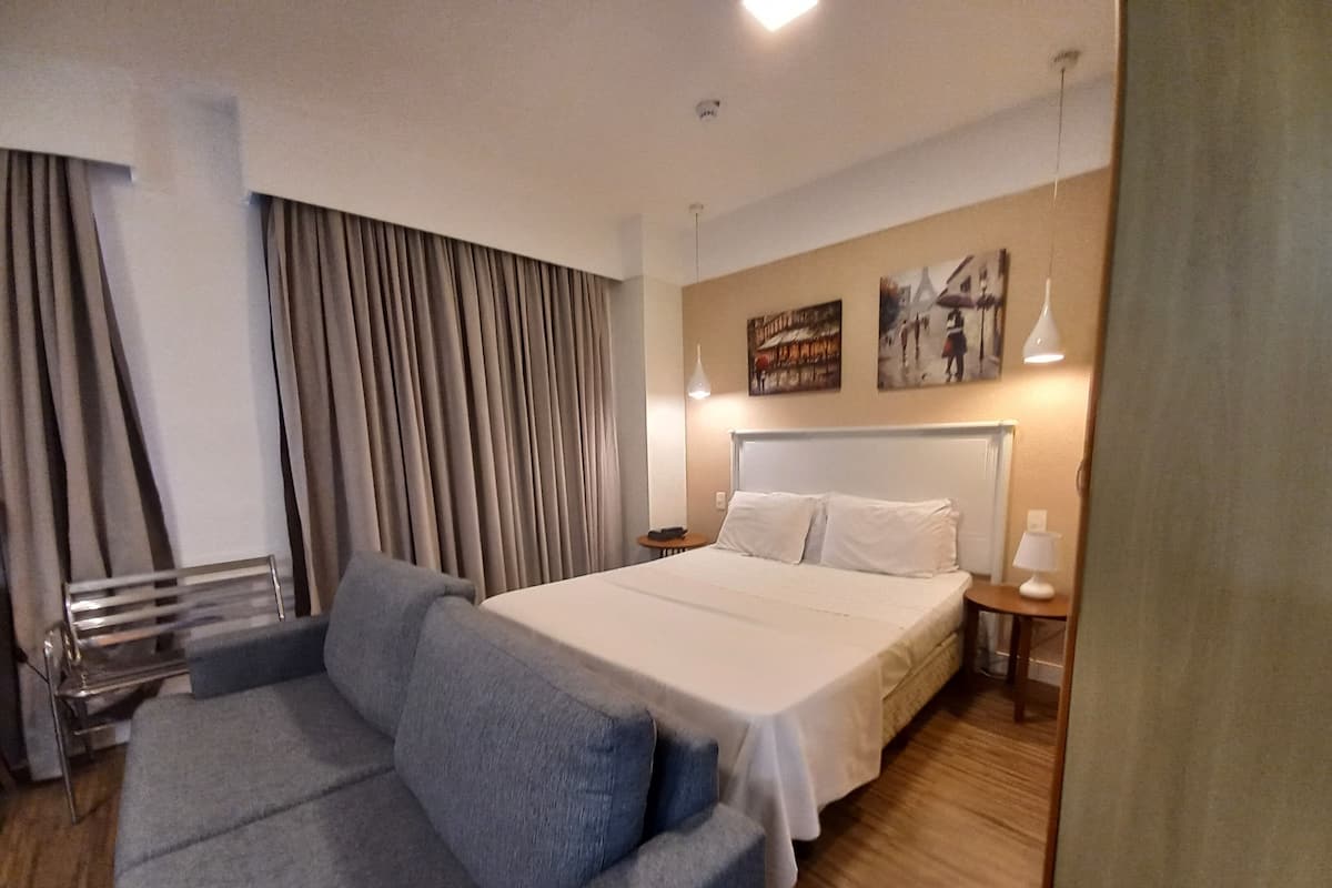Hotellihuone, jossa on suuri sänky, harmaa sohva, pieni pöytä ja seinään kiinnitetty lamppu.
