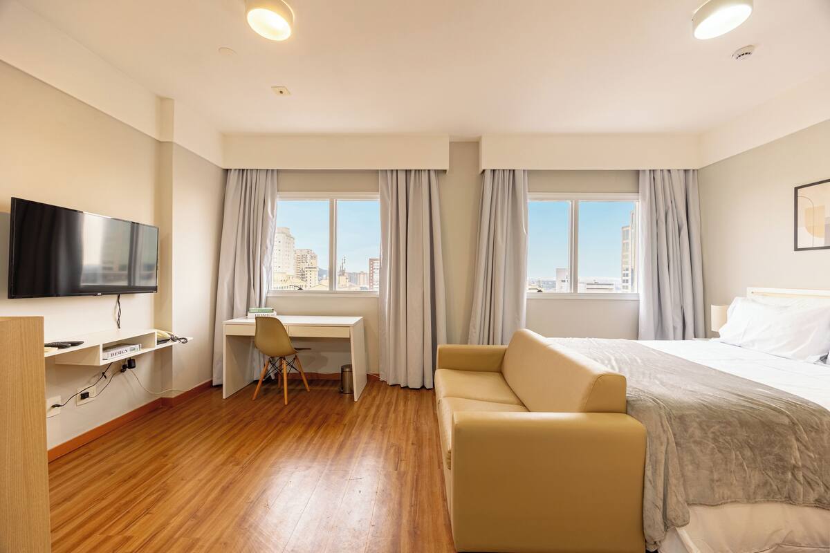 Hotellihuone, jossa on sänky, työpöytä, tuoli, televisio ja suuret ikkunat, joissa on verhot.
