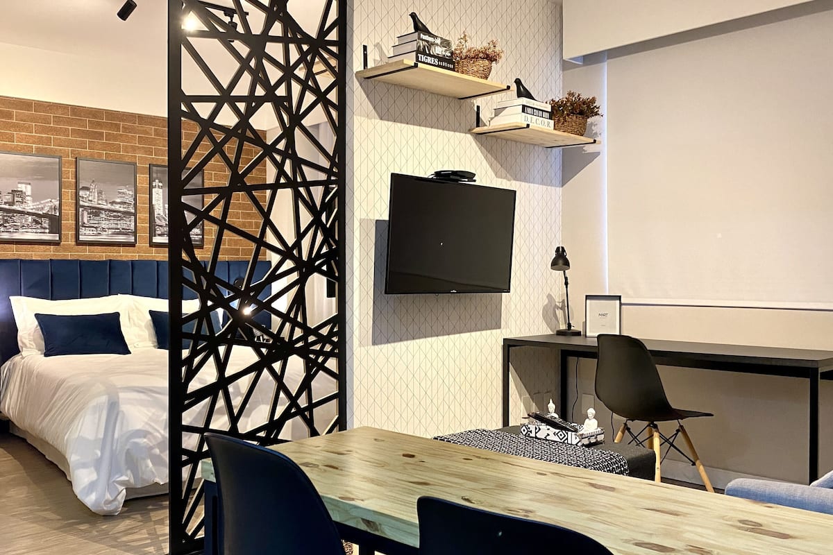 Moderni hotellihuone, jossa on sänky, työpöytä, tuoli, televisio ja hyllyjä, joilla on koriste-esineitä.