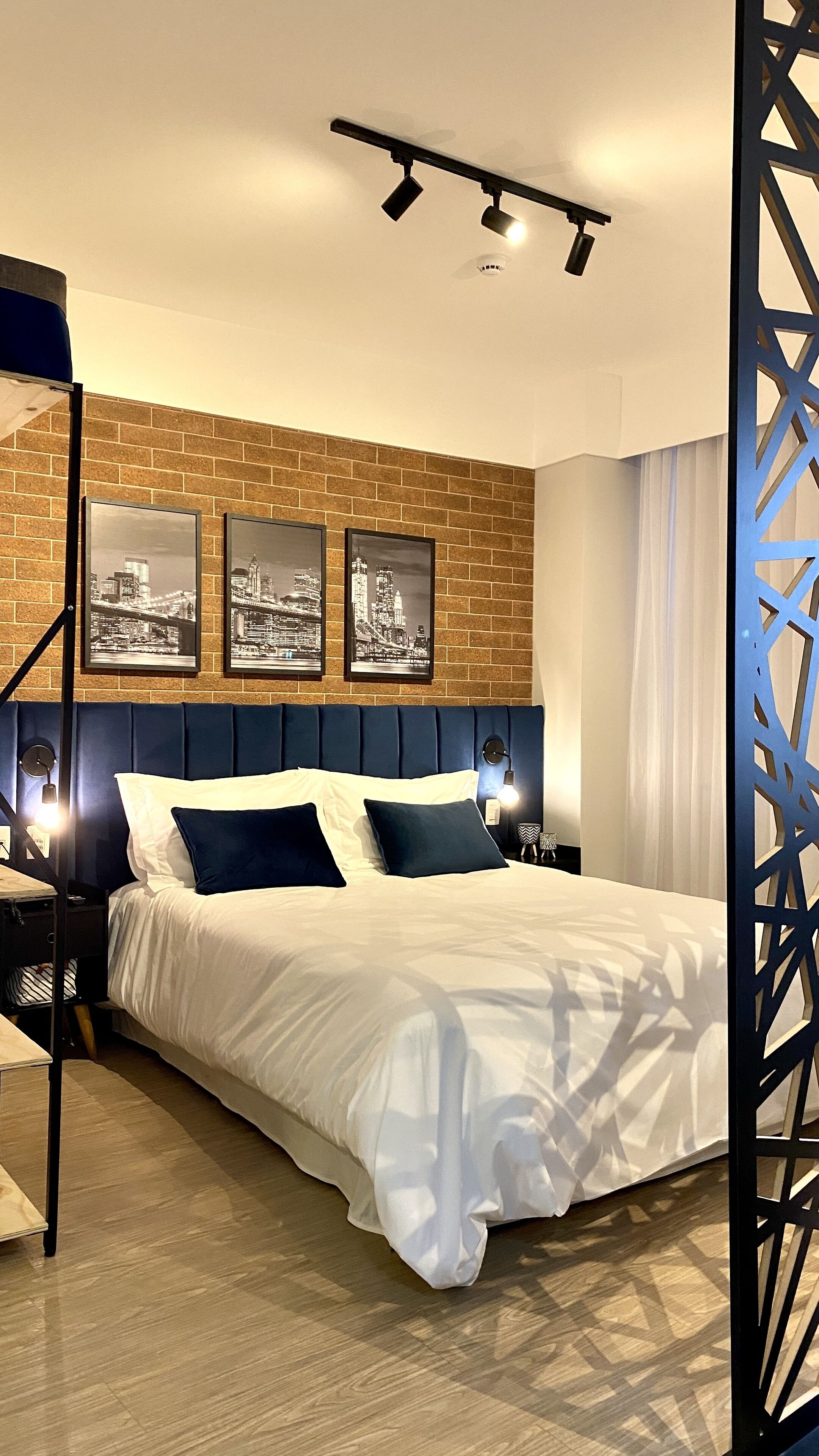 A modern hotel room with a brick-patterned wall, a large bed with white and dark blue bedding, and three black-and-white cityscape pictures.