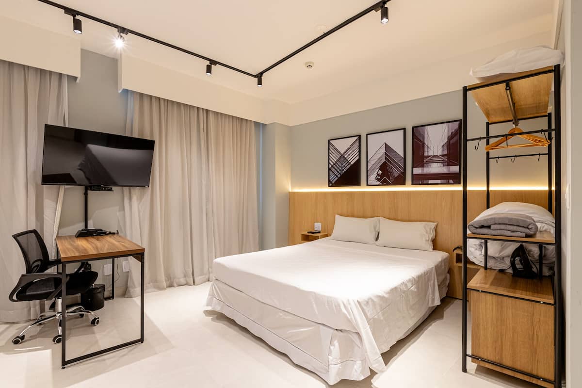 Moderni hotellihuone, jossa on suuri sänky, työpöytä tuolilla, televisio ja vaatekaappi hyllyineen.