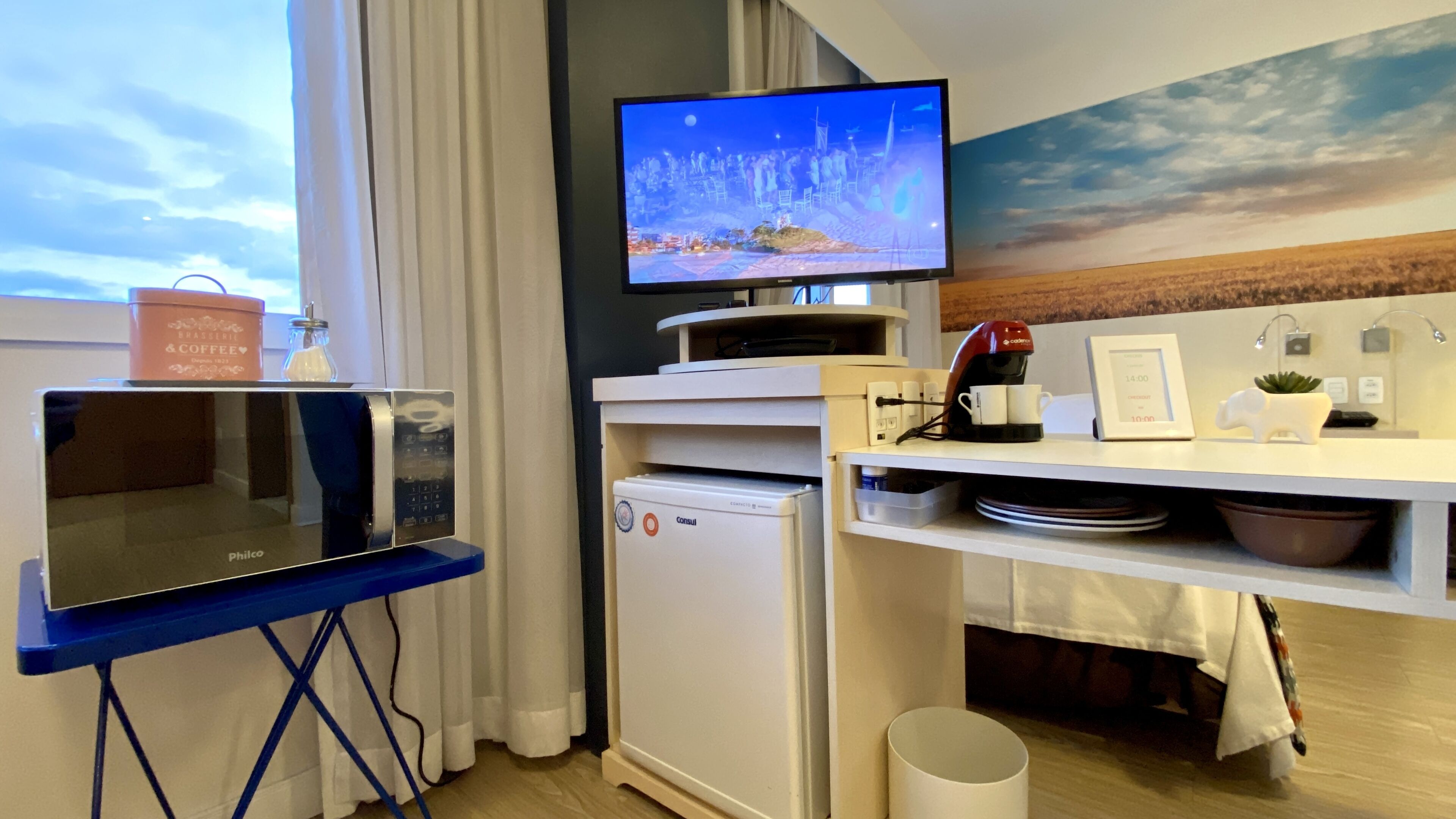 A compact hotel room with a kitchenette, a flat-screen TV, a coffee machine, and a small refrigerator.