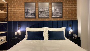 A hotel room with a brick wall, a bed with white and blue bedding, two bedside tables with lamps, and three framed cityscape pictures on the wall.