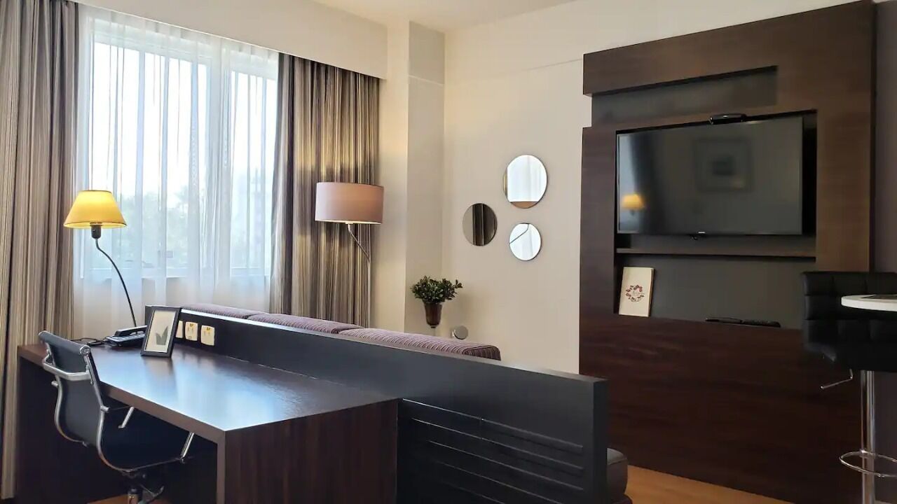 Une chambre d’hôtel moderne avec un bureau en bois, un canapé, un téléviseur fixé au mur et une grande fenêtre avec des rideaux.
