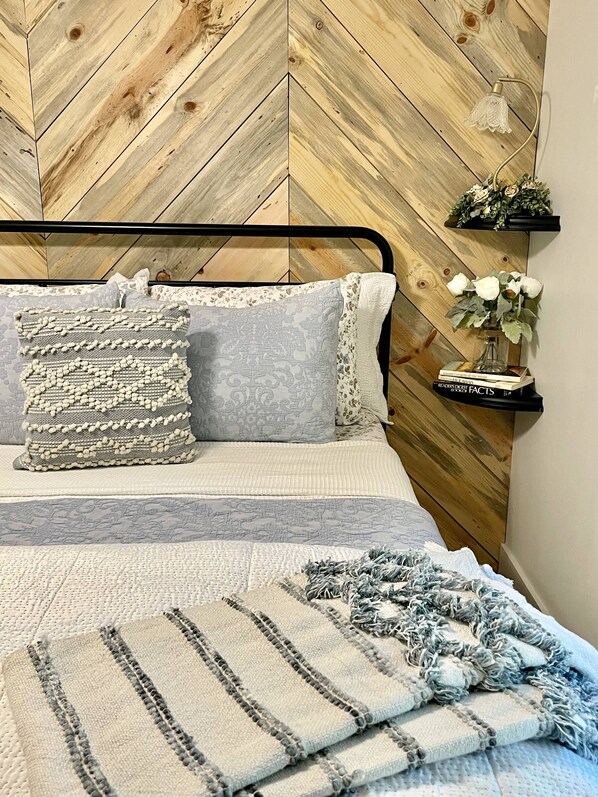 Un lit avec une tête de lit à motifs chevrons, un coussin décoratif, un plaid rayé et une petite étagère sur laquelle est posé un vase de fleurs.